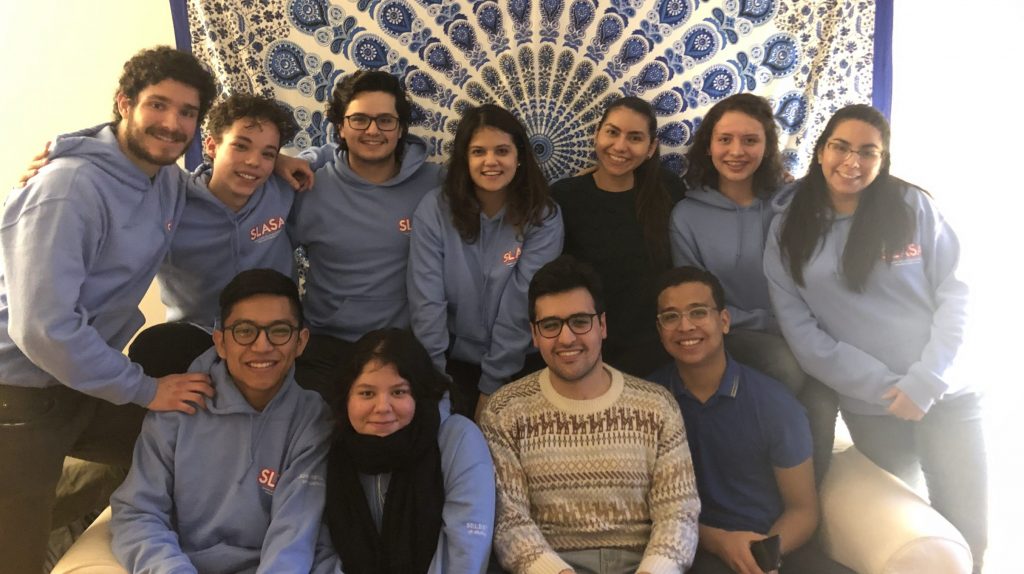 Image of 11 individuals in blue sweaters with the word "SLASA" posing in front of a fabric