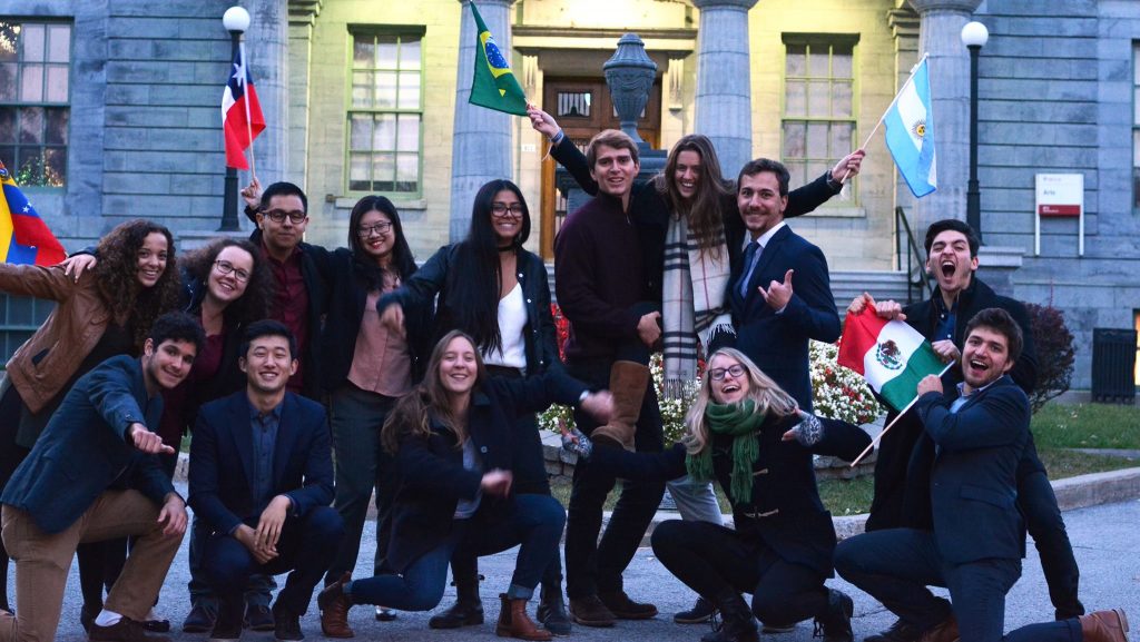 Image of various individuals laughing in front of an antique building holding flags from the country they represent